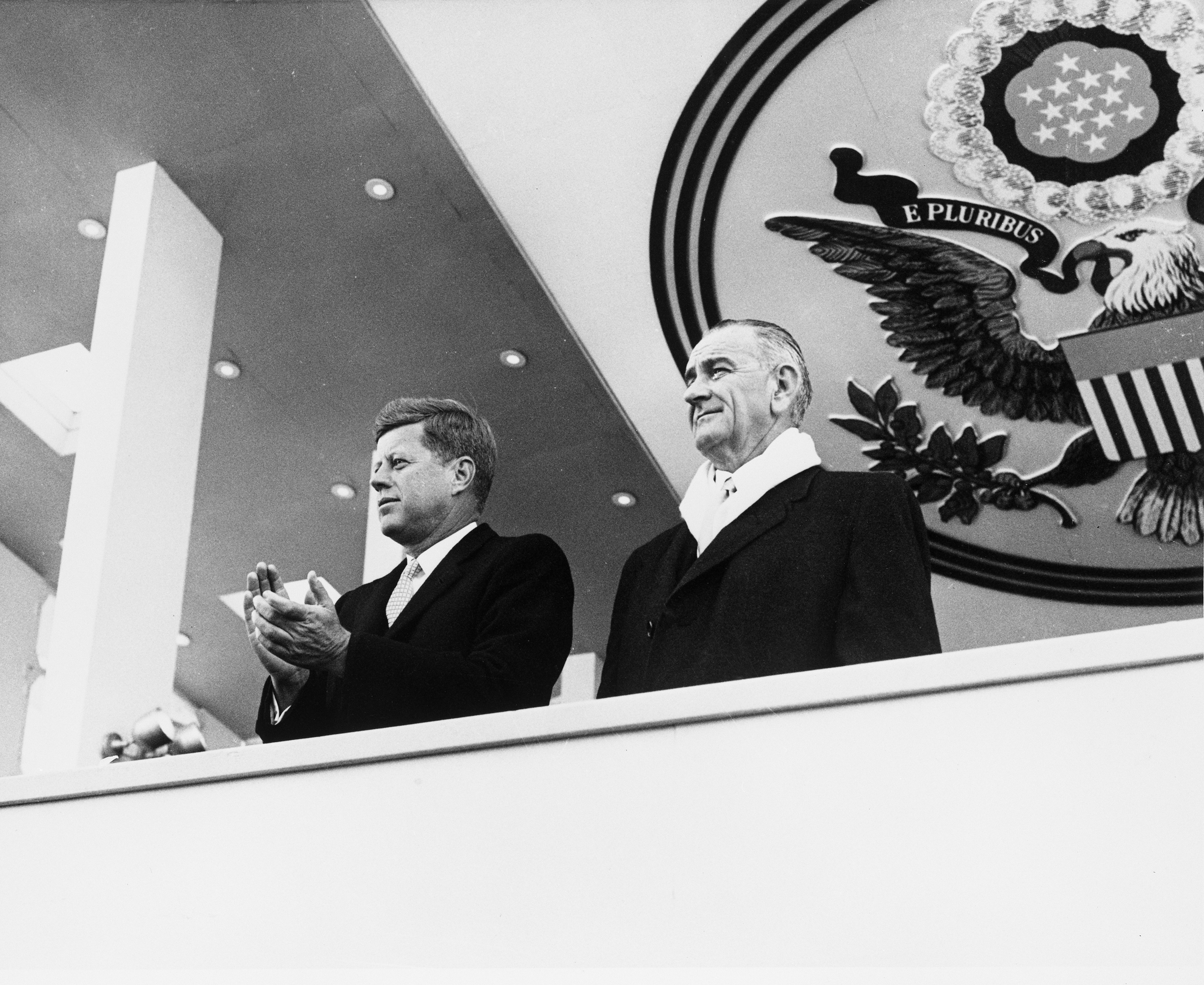President Kennedy and Vice President Johnson watch their inauguration parade from a balcony, with the United States seal behind and above them.